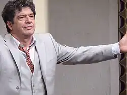 Un homme d'une cinquantaine d'années en costume gris, les bras ouverts.