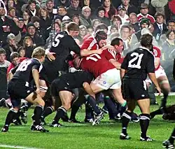 Hommes debouts dans un regroupement. Deux hommes en noir, au premier temps, surveillent cette action.