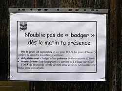 Gros plan d’une affichette de format A4 horizontal glissée dans une chemise plastique et agrafée à un panneau de bois. Dans un encadré gras, et sous le titre « N’oublie pas de « badger » dès le matin ta présence », l’avis annonce : « Dès le jeudi 21 septembre et ce pour tous les jours d’école (y compris le samedi), les enfants viendront : - obligatoirement « badger » leur présence dès leur arrivée à l’école, - éventuellement leur inscription à la cantine ou à l’étude surveillée. Tous les enfants de l’école devront donc avoir en permanence leur badge dans leur cartable. »