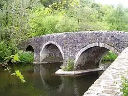 Photo d'un pont en pierres grises à trois arches enjambant une rivière bordée d'arbres feuillus