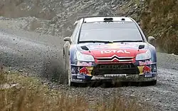 Citroën C4 WRC de Sébastien Loeb, livrée rouge et bleue, vue de face à pleine vitesse sur une piste boueuse.