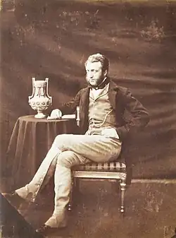 Portrait d'un homme assis sur une chaise à côté d'une table sur laquelle un vase est posé.