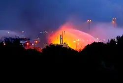 Phtographie couleur d'une zone industrielle fluviale en feu sur laquelle interviennent des pompiers avec des lances à eau.