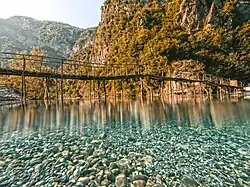 À travers une eau transparente, des cailloux bleus. Puis des reflets lumineux. Puis une passerelle fragile montée sur de hautes perches de bois ; puis une montagne arborée bouche la vue.