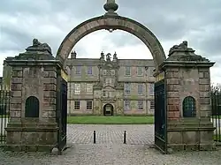 Au premier plan, porche d'entrée composé d'une voute en plein cintre appuyée sur deux loges en pierre. Les grilles grandes ouvertes ouvrent sur une cour pavée avec une pelouse centrale. Au fond, la façade du château, avec, au centre, le haut porche, orné, sculpté, surmonté d'une horloge, d'accès à la cour intérieure.