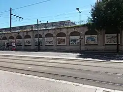 Photographie en couleurs. Des tableaux accrochés dans les arches d'un mur bordant une rue.