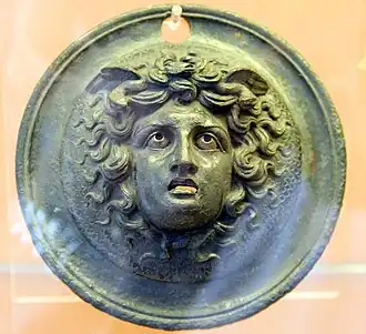 Dans un cercle, tête de Méduse entourée de cheveux, avec des serpents à la base du cou et des ailes en arrière de la tête. Bouche grande ouverte, yeux globuleux.