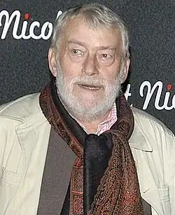 Photo couleur en buste d'un comédien d'une soixantaine d'année, il porte une chemise à carreaux, une écharpe et un trench gris.