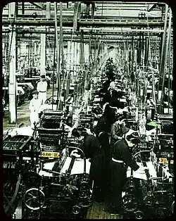Photo de l'intérieur d'une usine. Alignées à perte de vue, des machines sont en fonctionnement, contrôlées par des ouvrières.