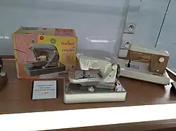 Deux machines à coudre dans une vitrine. Elles ont toutes les deux des coques en plastique clair. Celle de gauche, sans marque, a une teinte un peu argentée. Elle est exposée avec sa boîte d'origine, qui montre une photo de la machine sur un fond moitié rose, moitié jaune. À droite, la machine de marque Ma cousette est blanche, sauf la partie inférieure du bras et le centre de la tour qui sont bruns.