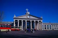 Photographie de face d'un bâtiment avec huit colonnes soutennant un toit en triangle. Des voyageurs se trouvent devant, ainsi que deux bus blancs et rouges sur le côté.