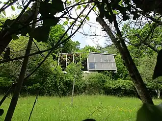 Photographie en couleurs en contre-plongée d’un bâtiment entouré de verdure. Des branches d'arbres au premier plan.
