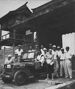 Photographie en noir et blanc d'une dizaine d'individus habillés en blanc autour d'une Jeep, devant un château.