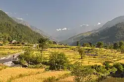 Vallée fertile avec des cultures en terrasses, quelques habitations et un passage à guet franchi par deux randonneurs, avec en arrière-plan des montagnes enneigées.