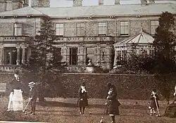 Photographie ancienne sur laquelle plusieurs enfants jouent au croquet dans le jardin à l'arrière d'un manoir avec une véranda imposante.