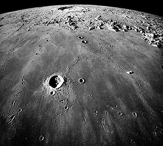 Image en plongée de la surface lunaire. Un petit cratère est visible au premier plan et un plus grand à l'arrière-plan.