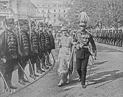 Une femme en grande tenue et un homme en uniforme de l'armée marchent dans une rue entourée de soldats.