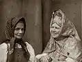 Photographie sépia d'une vieille femme portant un fichu, la main gauche serrée dans celles d'une quadragénaire portant une coiffe de tissu complexe avec motifs floraux, sur fond de rideau de théâtre.