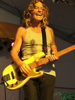 Mark Andes, holding his white bass and wearing a white T-Shirt, sings during a concert under an yellow spot.