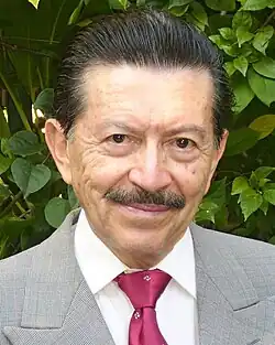 Homme brun moustachu de type sud-américain souriant en costume gris et cravate rose.