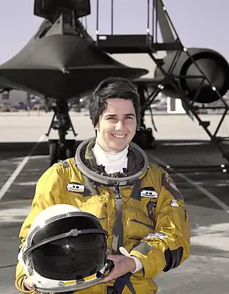 Marta Bohn-Meyer en combinaison d'aviatrice tenant un casque, devant un avion vraisemblablement un Lockheed SR671 Blackbird.