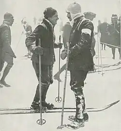 Photographie en noir et blanc d'une femme souriante et d'un homme moustachu à skis se faisant face.