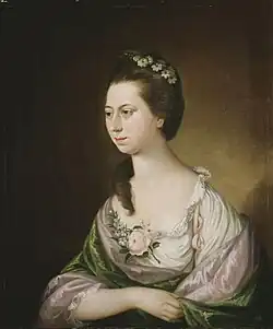 Portrait d'une femme adulte européenne, en trois quarts, avec des cheveux bruns et une couronne de fleurs blanches. Elle porte une robe crème avec des détails en dentelle, ornée d'une rose pâle.