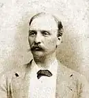 Photo en noir et blanc du torse et du visage d'un homme moustachu.