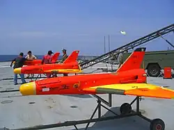 Trois drones rouges fuselés avec rampe sur pont de porte-avion. Les ailes delta et les nez sont jaunes (3&nbsp;×&nbsp;2,5&nbsp;m env.).