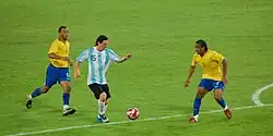 Photographie couleur. Au premier plan, Messi joue pour l’Argentine à l’entrée de la surface de réparation. Ses adversaires lui disputant la balle sont au nombre de deux et revêtent la tenue de l’équipe du Brésil de football.