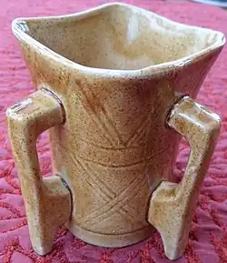 Photo d'un récipient en céramique émaillée de teinte blanc orangé, à quatre poignées de prolongeant par des pieds à la base.