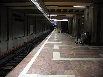 Une partie du quai central, le sol est en marbre rouge avec des motifs géométriques blancs, et une voie du métro