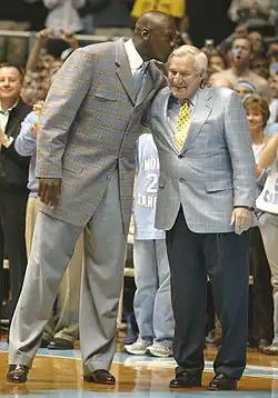 Michael Jordan en costume gris et cravate embrassant affectueusement le crâne de son ancien coach, Dean Smith, sur un parquet de basket-ball