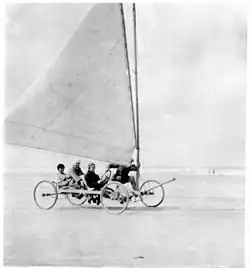 Photographie noir et blanc de quatre personnes à bord d'un char à voile sur une plage.