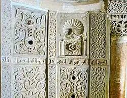 Photographie montrant quelques-uns des panneaux en marbre blanc, sculptés et ajourés, de la paroi du mihrab. Entre les panneaux est visible une partie de l'inscription coranique en caractères coufiques.