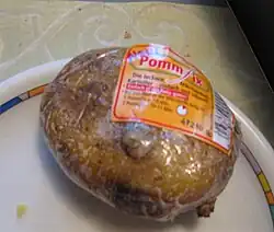 Posée sur une assiette, une grosse pomme de terre emballée d’un plastique transparent présente son étiquette orange avec les mentions de marque et temps de cuisson, et un code barre blanc et noir.