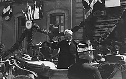 Photo en noir et blanc d'un homme aux cheveux blancs, portant un nœud papillon et un costume trois pièces, en train de saluer la foule avec son chapeau ; en arrière-plan, un bâtiment de la République française est visible