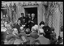 Photographie noir et blanc d'un homme moustachu lisant un discours à l'entrée d'une maison, devant un public d'hommes et de femmes.