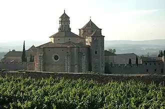 La photographie couleur représente une vigne en période végétative devant un mur d'enceinte. En arrière-plan apparaîtl'église du monastère de Poblet.