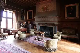 Vue d'une pièce meublée, fauteuils autour d'une table de jeu, piano dans un angle.