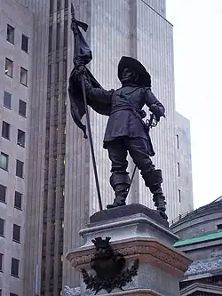 Statue de Maisonneuve portant fièrement un drapeau de la main droite et une épée à la main gauche. Il porte un large chapeau au bord relevé, une tunique, des pantalons et de hautes bottes aux bords repliés.