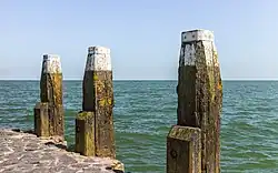 Trois poutres jaillissent du bord d'un quai. Leur bout dans le ciel est souligné de blanc. Derrière, la mer.