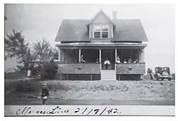 Une photographie en noir et blanc d'une maison à un étage et demie, avec une galerie en avant sur laquelle se tiennent 7 personnes. À droite de la maison, une voiture sombre est stationnée. En avant plan, un enfant aux cheveux clairs se tient sur la route. À gauche de la maison, un bosquet.