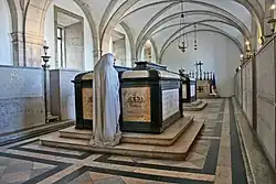 Sépultures du roi et de la reine de Portugal dans une crypte voûtée, les monuments funéraires sont en pierres claires et encadrées d'une armature noire, ils portent deux couronnes sculptées et font face à une statue blanche de pleureuse