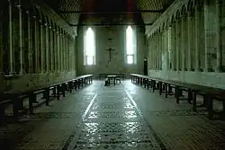 Le long des murs rythmés par des colonnades, d’une longue et haute salle, chichement éclairée par deux hautes baies deux rangées de tablées et bancs se font face. Au fond, sous une grande croix, se trouve placée perpendiculairement la table réservée aux supérieurs.