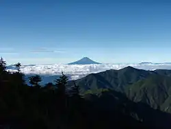 Photo couleur de la silhouette du sommet conique d'une montagne, émergeant d'une mer de nuages blancs au loin (centre de la photo). Un massif montagneux boisé, au premier plan et un ciel bleu en arrière-plan.