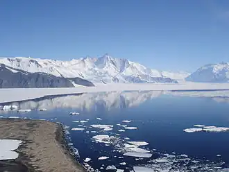 Montagne acérée enneigée devant une baie reflétant un ciel bleu.