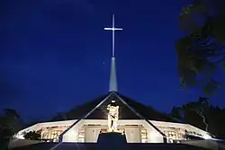 Photo nocturne d'une église en forme de cône large et aplati, surmontée d'une haute croix, derrière une statue représentant André Kaggwa mutilé.