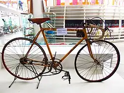 Photographie d'un vélo ancien au cadre jaune placé sur un présentoir, une étiquette blanche apposée sur le cadre.