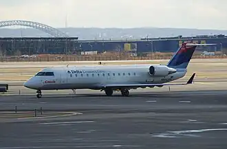 Photo de trois quart face d'un avion de ligne, aux couleurs blanches avec des inscriptions bleues et rouges, à la porte d'embarquement d'un aéroport avec la passerelle passagers contre la porte opposée de l'avion.